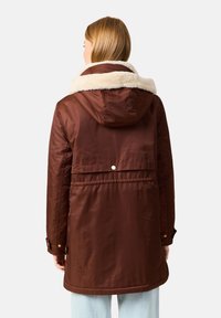 Ruskea, pehmustettu takki, jossa on turkisvuorattu huppu ja pidempi leikkaus. Takissa on nappikiinnitys selässä ja kultaiset yksityiskohdat hihansuissa.