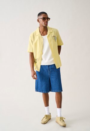 Jeune homme portant une chemise jaune à manches courtes, un débardeur blanc, un short en jean, des chaussures beiges, des chaussettes blanches et des lunettes de soleil, debout devant un fond uni.