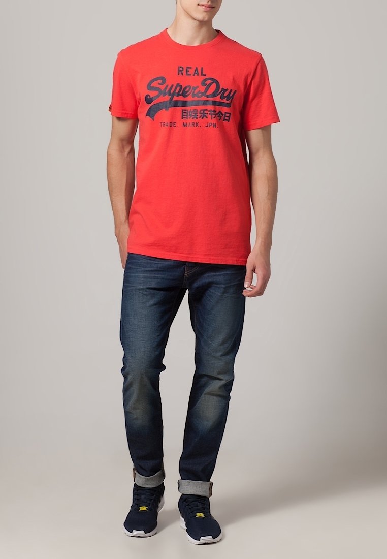Camiseta de algodón roja con texto negro, combinada con jeans de mezclilla azul. El modelo lleva zapatillas marinas con suelas blancas. Diseño simple, ajuste relajado.