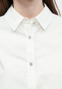 Λευκό βαμβακερό πουκάμισο με κλασικό γιακά και πέντε λαμπερά κουμπιά με διακοσμητικά στοιχεία. Λεία υφή με καθαρό, απλό σχέδιο.