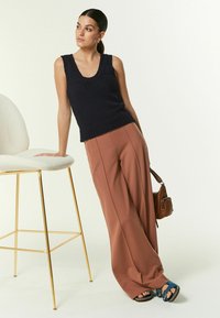 Top sans manches en maille bleu marine associé à un pantalon brun à taille haute et jambes larges, accompagné de sandales bleues à brides, placé à côté d'une chaise claire.