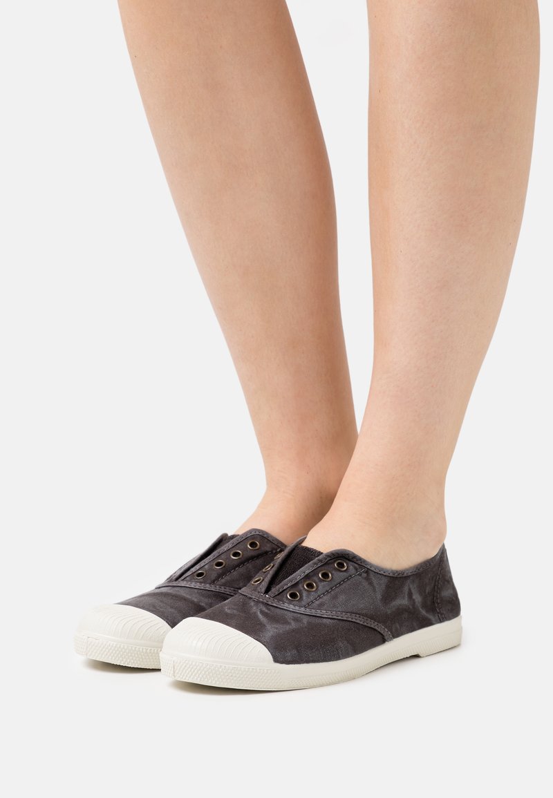Ballerines en toile grises sans lacets avec un embout et des œillets en caoutchouc blanc, présentant une surface texturée, reposant sur des chevilles nues.