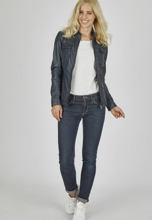 Dunkle Lederjacke mit Reißverschluss, ausgestattet mit zwei Fronttaschen; kombiniert mit schmal geschnittenen blauen Jeans und weißen Sneakers. Das Modell steht vor einem neutralen Hintergrund.