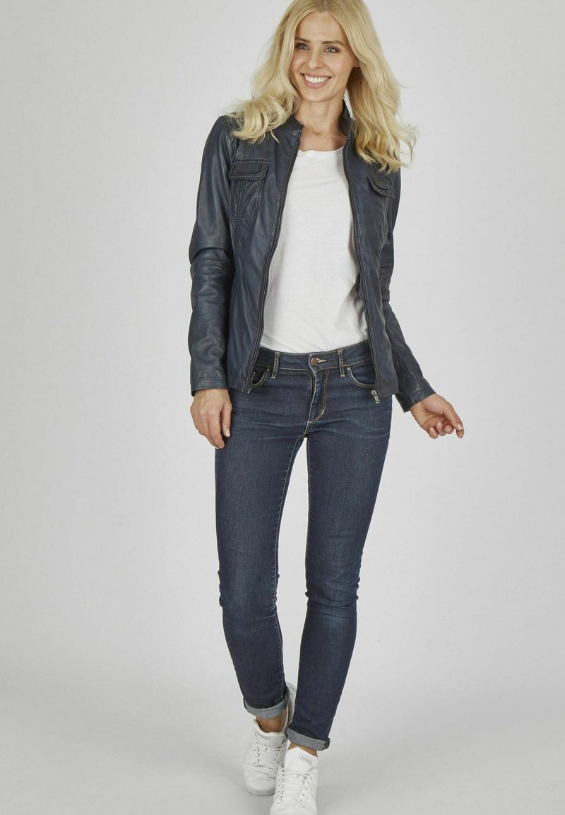 Veste en cuir noir avec une fermeture éclair, dotée de deux poches avant ; associée à un jean bleu ajusté et des baskets blanches. Le modèle se tient sur un fond neutre.