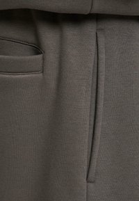 Tessuto verde scuro, texture visibile che mostra i dettagli di una tasca e cuciture senza soluzione di continuità. Il materiale appare morbido e leggermente elastico.