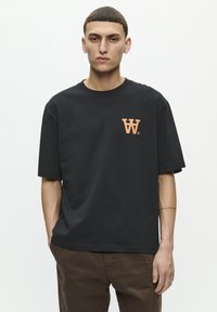 Zwarte katoenen T-shirt met een losse pasvorm, korte mouwen en een ronde hals. Bevat een oranje logo op de linker borst.