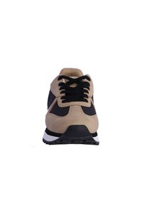 Zapatilla beige y negra con materiales de ante y malla, que presenta una punta redondeada, cordones negros, suela blanca y suela exterior de goma texturizada.