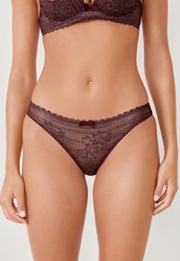 Perizoma in pizzo bordeaux con motivi floreali, bordi a scallop e una piccola decorazione a fiocco nella parte anteriore della vita. Texture morbida e trasparente.