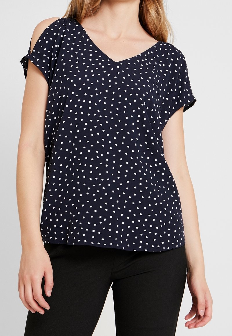 Femme portant un chemisier bleu marine à manches courtes avec des pois blancs et un pantalon noir, vue du cou aux hanches.