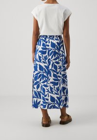 Henkilö nähtynä takaa, päällään valkoinen lyhythihainen paita, sinivalkoinen kuvioitu midi-hame ja ruskeat sandaalit yksinkertaisella taustalla.