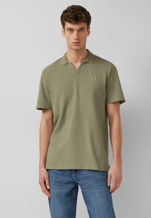 Młody mężczyzna stojący, ubrany w oliwkowozieloną koszulkę polo z krótkim rękawem i kołnierzykiem oraz niebieskie dżinsy, na jednolitym szarym tle.