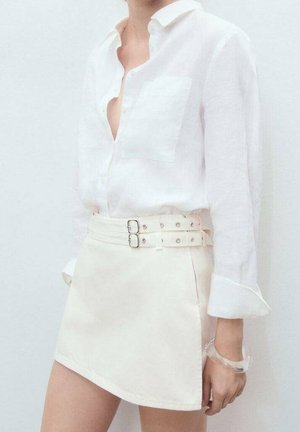 Personne portant une chemise blanche boutonnée avec des manches retroussées et une mini-jupe blanche avec un détail de double boucle à la taille.