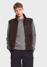 Jeune homme portant une veste polaire marron zippée sur un pull gris et un pantalon cargo noir, debout les mains dans les poches sur un fond uni.