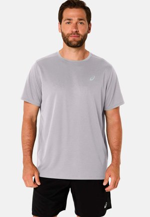 Man die een lichtgrijs sportshirt met korte mouwen en een zwarte korte broek draagt, staand tegen een effen witte achtergrond.