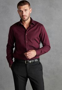 Burgunderfarbene Langarm-Hemd mit subtiler Struktur, kombiniert mit schwarzen Hosen und einem eleganten schwarzen Gürtel, der eine maßgeschneiderte Passform zeigt.