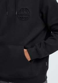 Juodas džemperis, pagamintas iš minkštos medžiagos, su siuvinėta žemėlapio logotipu krūtinėje, priekinė kišenė ir įaustais rankogaliais bei apačia.