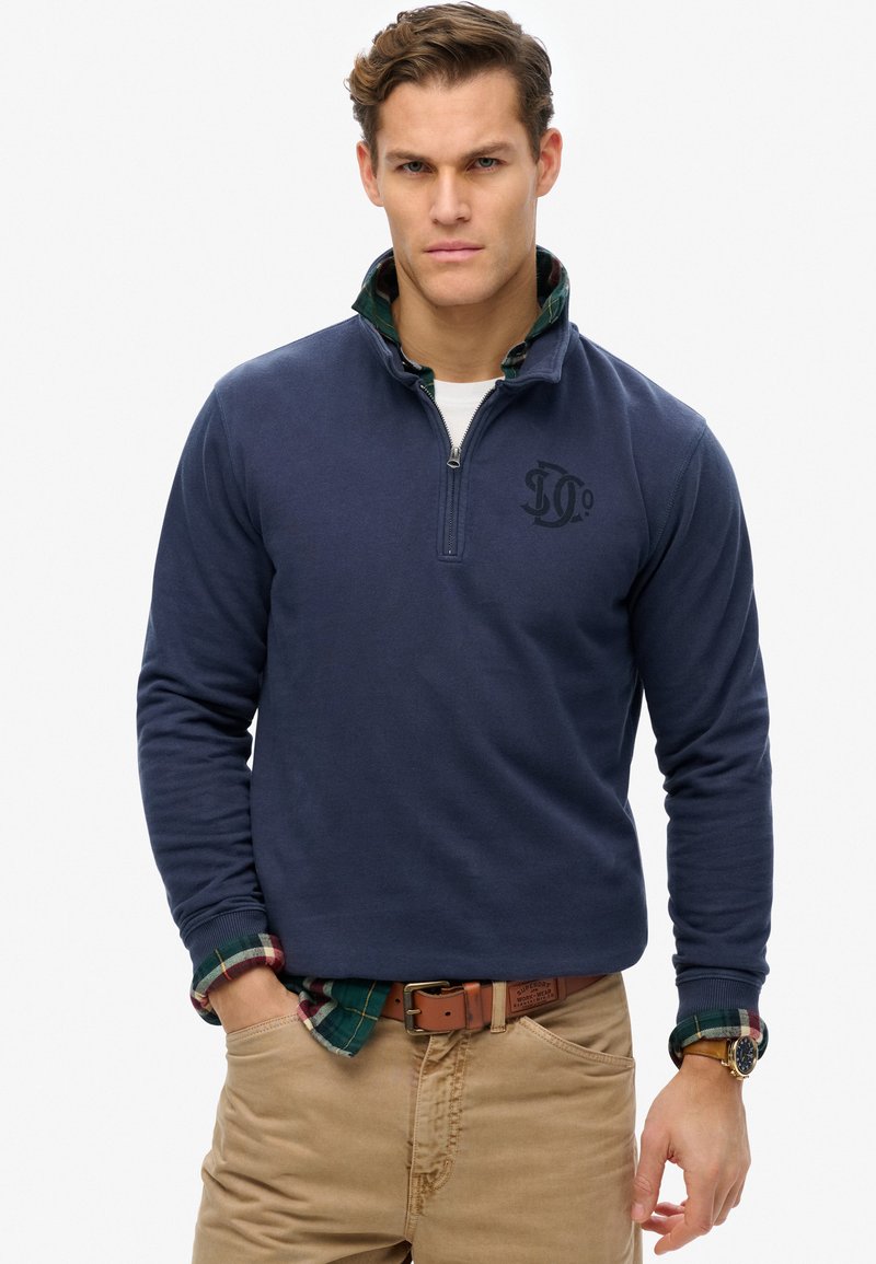 Sudadera de forro polar azul marino con cremallera de un cuarto, presenta un logo en el pecho, llevada sobre una camisa verde a cuadros, combinada con pantalones caqui.
