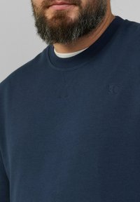 Sweat-shirt bleu marine avec un col rond, en tissu texturé et un logo subtil sur la poitrine. Présente des coutures renforcées aux joints.