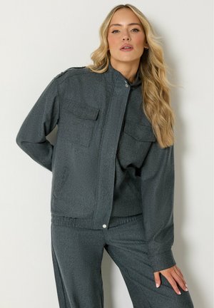 Donna con i capelli biondi e lunghi che indossa una giacca grigio scuro abbottonata e pantaloni abbinati, posando con una mano sul fianco su uno sfondo semplice.