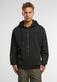 Felpa nera con zip realizzata in un materiale morbido, dotata di tasca frontale, cordini regolabili e dettaglio con zip sulla manica.