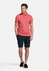 Rode polo shirt met witte afwerking, gecombineerd met donkere shorts. Model draagt zwarte instappers. Eenvoudig, casual ontwerp en aansluitende pasvorm.