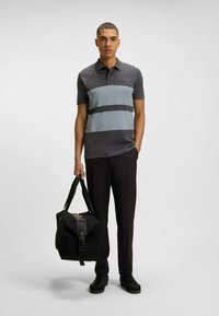 Koszulka polo z ciemnoszarymi, jasnoszarymi i czarnymi poziomymi paskami. Model trzyma czarną torbę, ma na sobie czarne spodnie i buty.