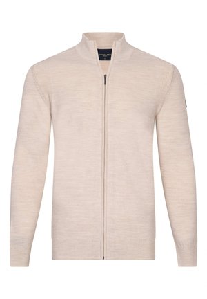 Mann trägt einen beigefarbenen Reißverschluss-Pullover über einem gemusterten Hemd, die Arme verschränkt und eine Hand nahe am Gesicht erhoben.