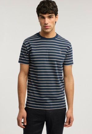 Junger Mann mit welligem dunklem Haar, der ein kurzärmeliges T-Shirt mit horizontalen Streifen in Marineblau und Weiß sowie schwarze Hosen trägt und vor einem einfarbigen Hintergrund steht.