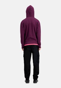 Sweat à capuche violet avec capuche, porté sur un t-shirt rose, associé à un pantalon cargo noir. La tenue présente un design décontracté et une coupe ample.
