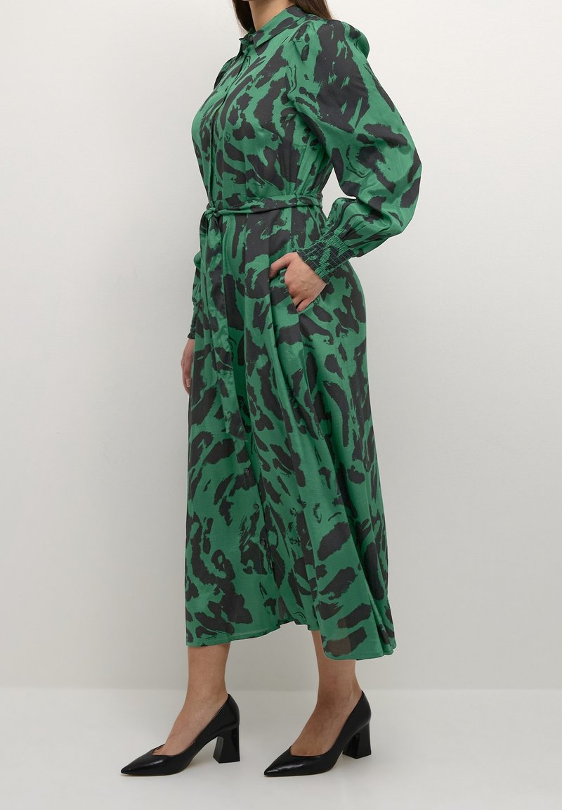Femme portant une robe midi à manches longues à motifs verts et noirs avec une ceinture à la taille et des chaussures noires à talons carrés, debout de profil.