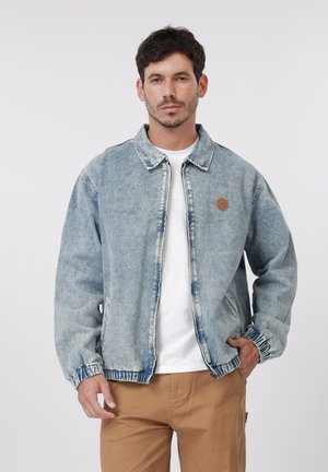 Moški, oblečen v svetlo modro denim jakno čez belo majico in rjave hlače, stoji z eno roko v žepu na enobarvnem ozadju.