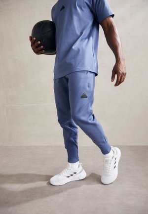 Conjunto deportivo azul que incluye una camiseta de manga corta y pantalones joggers a juego, combinado con zapatillas blancas. Bola negra sostenida en la mano derecha.