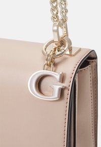 Beige läderhandväska med en guldkedjeslip, med en stor silverbokstav "G" charm och minimal sömnad längs kanterna.