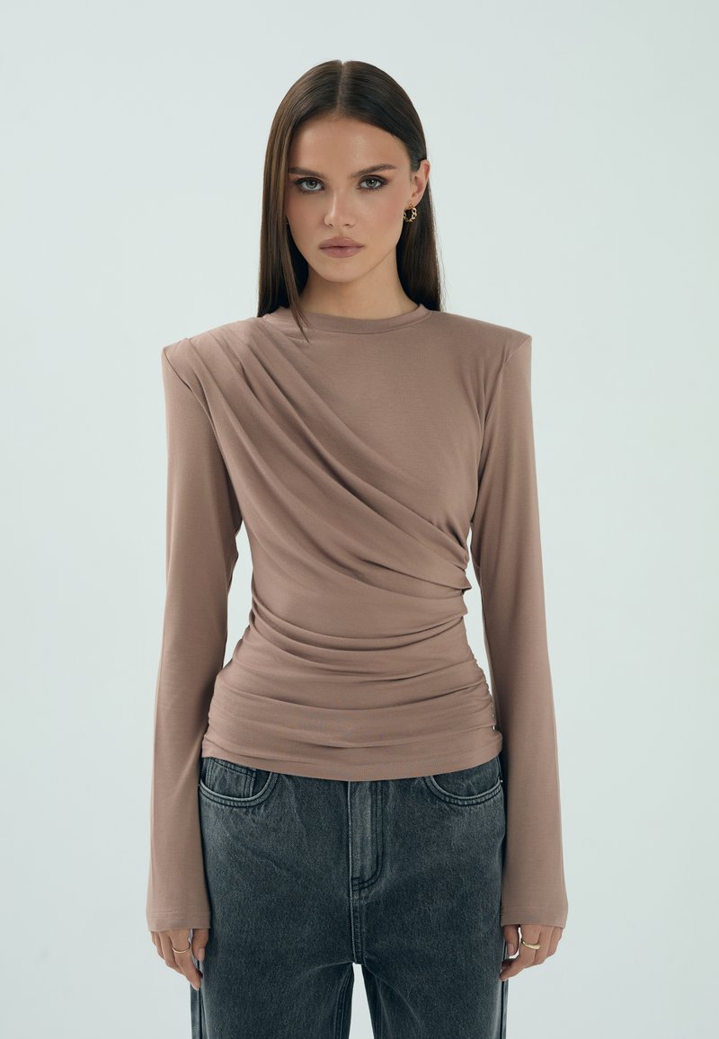 Długi rękaw, dopasowana bluzka w odcieniu taupe z falbaniastym wzorem i gładką fakturą. Połączona z wysokim stanem ciemnoszarych dżinsów.