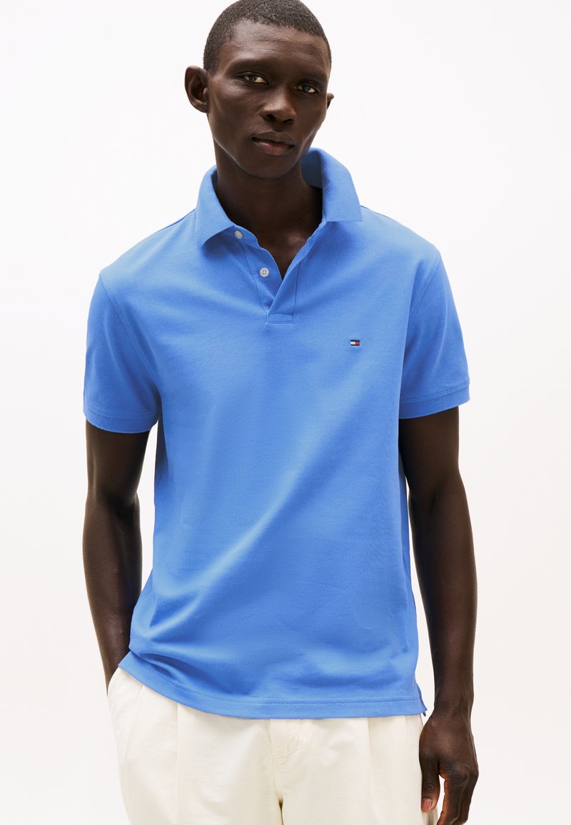 Mand iført en lyseblå kortærmet poloshirt med et lille logo på brystet og hvide bukser, stående mod en ensfarvet baggrund.