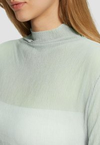 Lichtgroene top met hoge hals, kenmerkend door een textuur en ribbelontwerp met een verloopeffect. Zacht stof en lichtdoorzichtige delen.