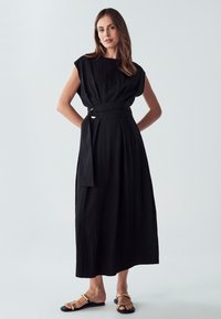 Črna brezrokavna obleka s tekočo silhueto, značitnim pasom in strukturirano tkanino. Ujemajoči se soflatni sandali z zlatimi detajli.