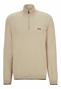 Beige stickad tröja med halvdragkedja, med en svart logotyp på bröstet och svarta detaljer vid midjan och ärmarna.