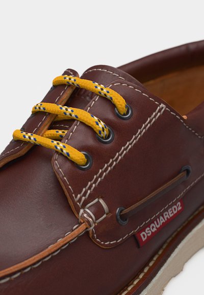 Chaussure bateau en cuir marron avec des lacets à motifs jaunes et bleus, une couture blanche, et une étiquette rouge DSQUARED2 sur le côté.