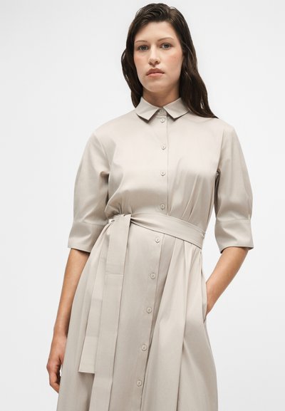 Femme aux longs cheveux foncés portant une robe beige boutonnée avec un col, des manches trois-quarts et une ceinture nouée, debout devant un fond uni.
