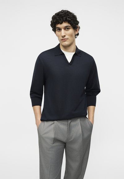 Νέος άνδρας με σγουρά μαλλιά, φοράει μαύρο πουκάμισο με μακριά μανίκια πάνω από λευκό εσώρουχο και γκρι πλισέ παντελόνια, με τα χέρια στις τσέπες.