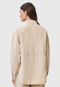 Camisa beige de botones con un corte relajado, que presenta mangas largas, un cuello y un detalle de costura en la parte posterior. Textura suave.