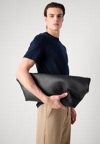 Juodas odinis krepšys su švelnia, lygiomis tekstūra, stačiakampio formos ir minimalistiniu dizainu, laikomas po pažastimi, derinamas su tamsiai mėlynu marškinėliu.