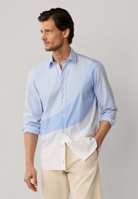 Man draagt een blauw-wit gestreept shirt met lange mouwen en een gebogen patroon, beige broek, met zijn linkerhand in zijn zak.