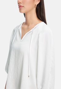 Weißer Kapuzenpullover mit V-Ausschnitt, Kordelzug und weit geschnittenen Ärmeln, aus leichtem Stoff mit glatter Textur.