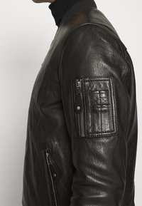 Schwarze Lederbomberjacke mit einem gerippten Kragen. Verfügt über eine Reißverschlusstasche am linken Ärmel und strukturierten Details an der Schulter.