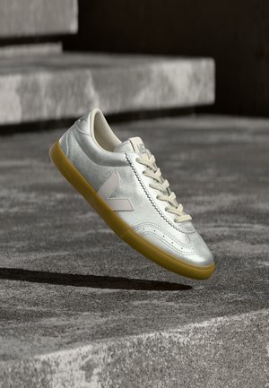 Silberner Sneaker aus veganem Leder mit abgerundeter Zehenpartie, beigen Schnürsenkeln und gelber Gummisohle. Mit einem weißen Logoakzent an der Seite.