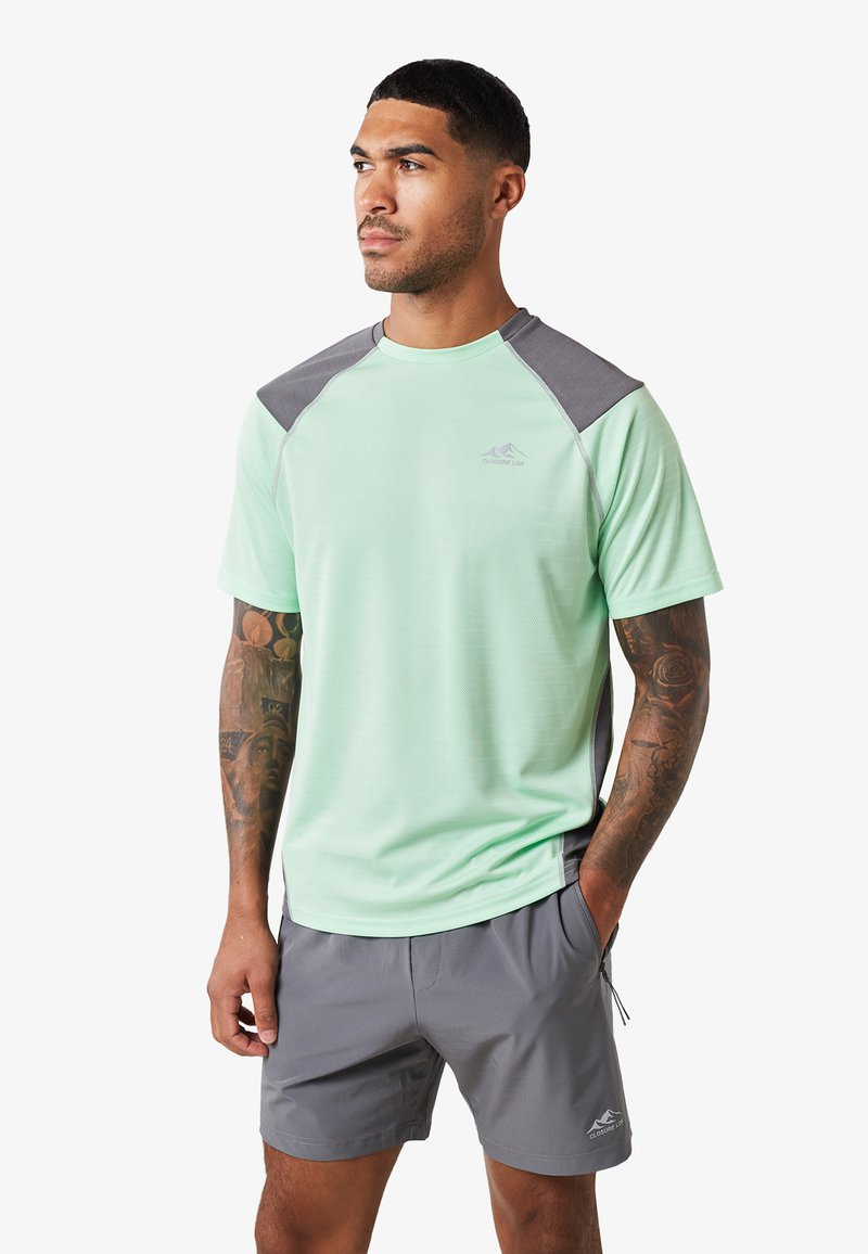 Camisa deportiva de manga corta verde claro con detalles grises en los hombros, cuello redondo y una textura sutil. Se lleva con pantalones cortos grises.
