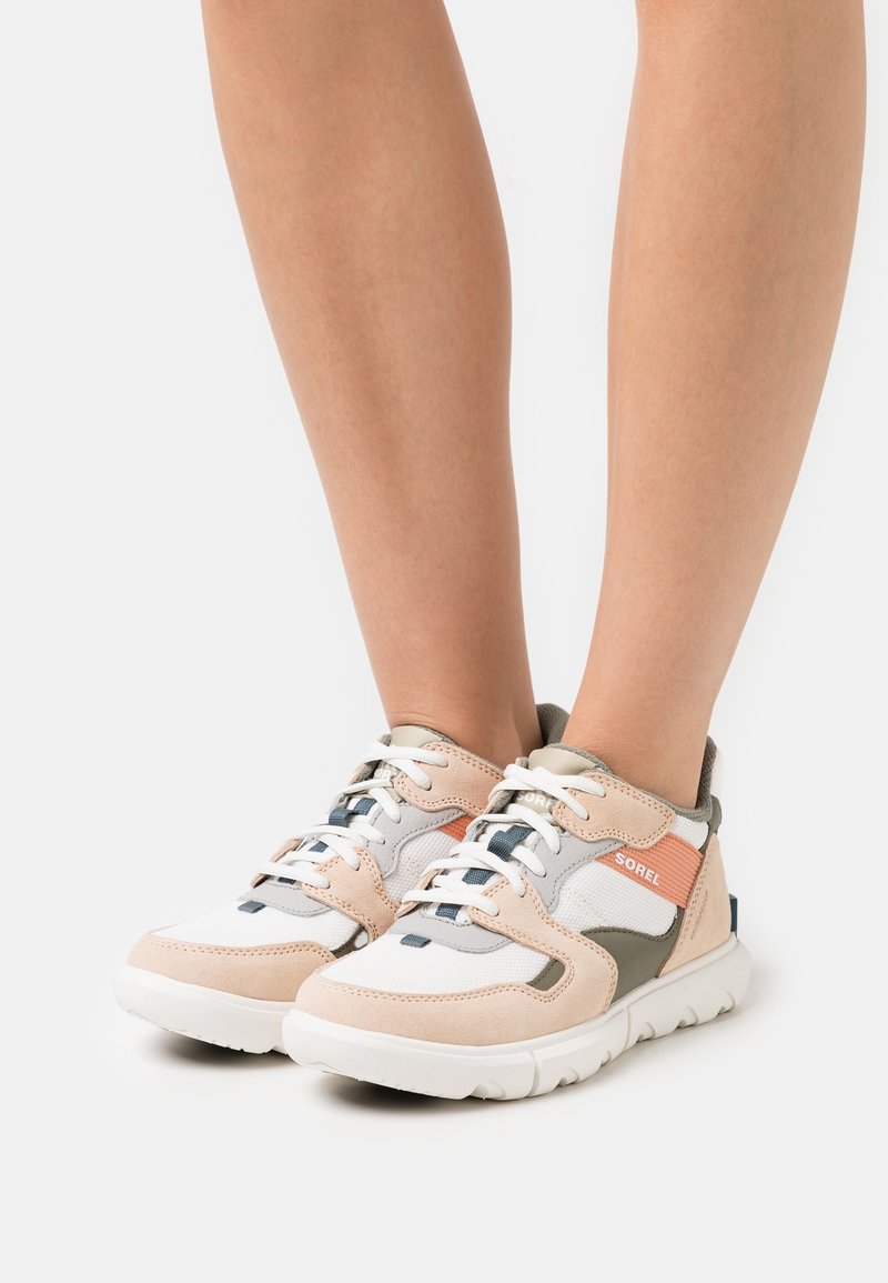 Jambes portant des baskets basses à lacets en blanc, beige, gris et orange avec des semelles épaisses blanches sur un fond blanc.