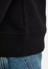 Zwarte sweatstof, geribbelde zoom, zijnaaddetail met klein zwart logo-label, gecombineerd met vervaagde blauwe denim. Eenvoudig, strak ontwerp.
